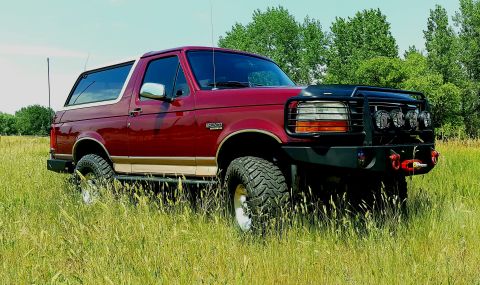1996 Ford Bronco - Burgundy Ford Bronco Eddie Bauer with tan two-tone lower body, photographed in 3/4 front view standing in tall summer grass. It wears a bla…