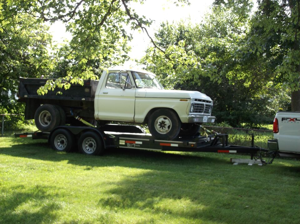 White 1970s dump truck on a trailer towed by a white pickup truck.