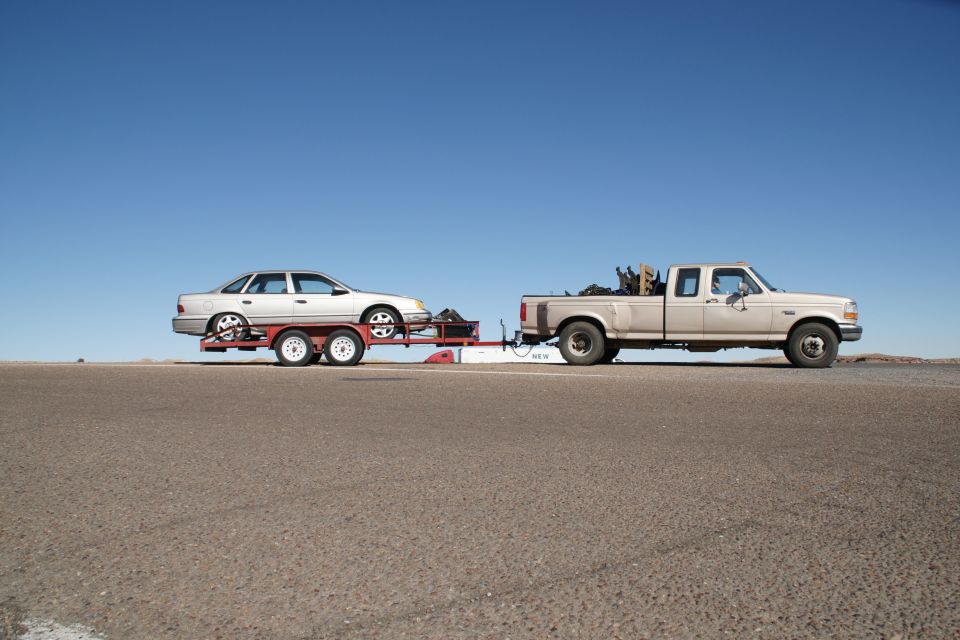 Light-colored sedan on a trailer towed by a pickup truck.