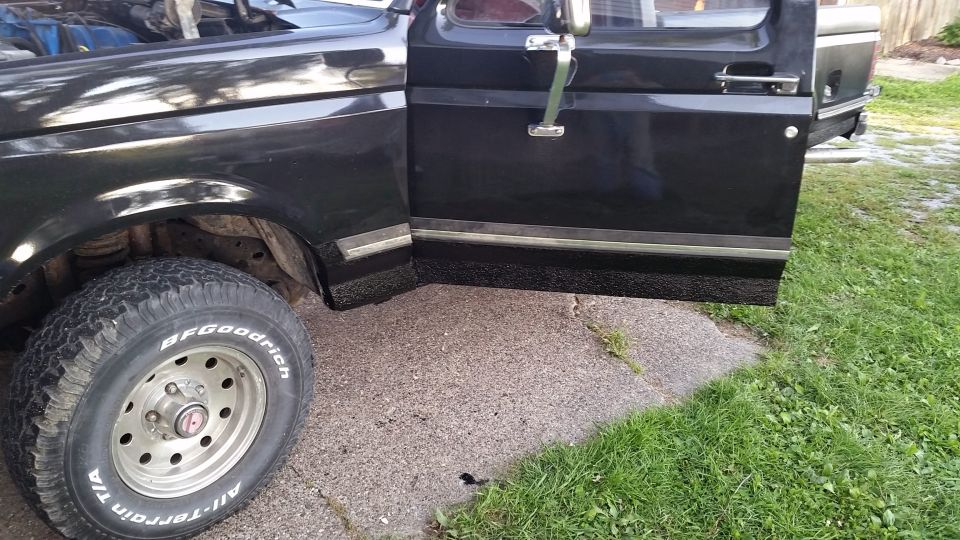 Black Ford pickup truck driver's side door and fender with open door, showing textured lower body and front tire.