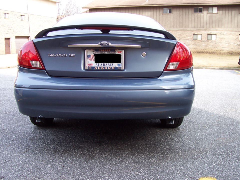 Rear view of a blue-gray Ford Taurus SE sedan with spoiler and Alabama Auburn plate, parked by brick buildings