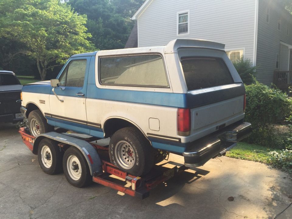 Blue and white Ford Bronco SUV on a red tandem-axle trailer in a driveway, rear driver-side view