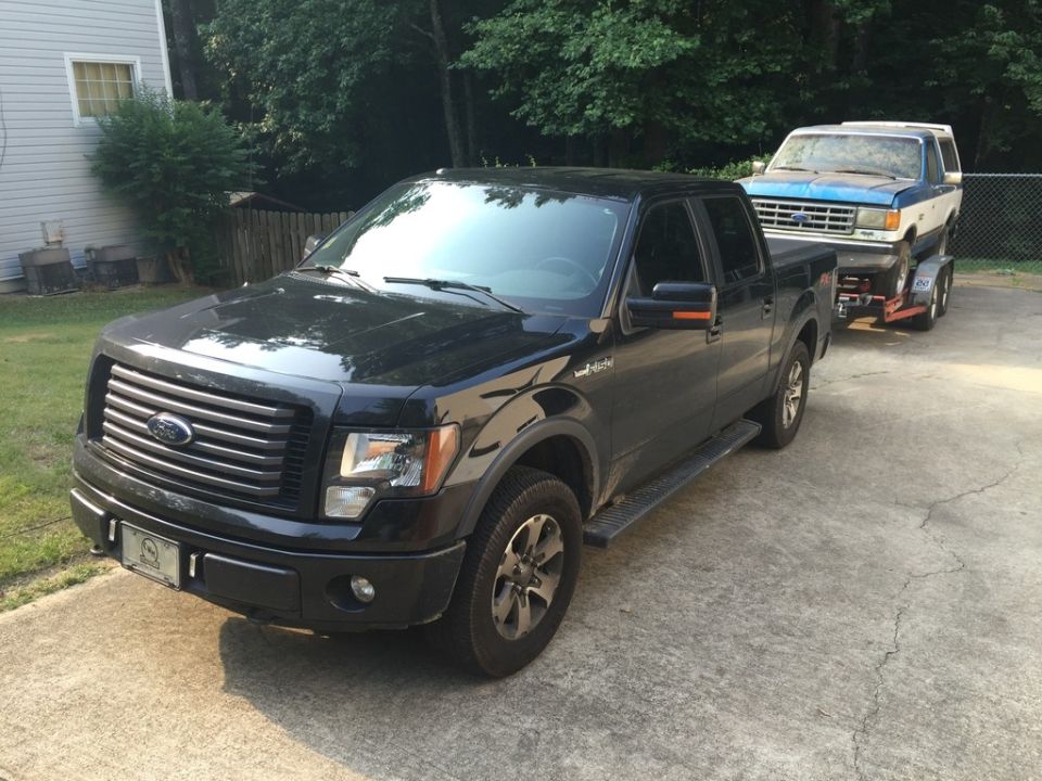Black Ford F-150 towing a faded blue and white Ford Bronco on a trailer in a driveway