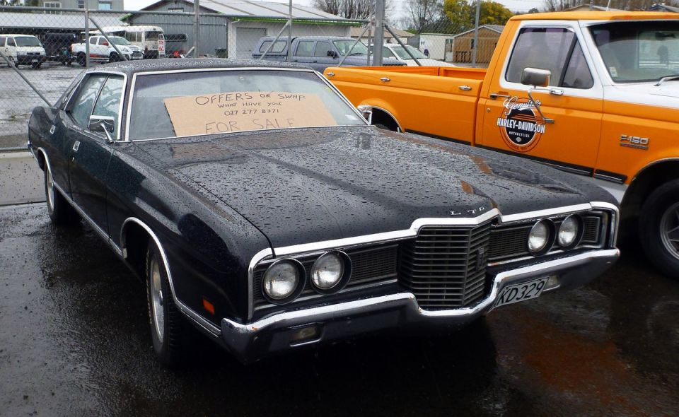 Dark sedan with "LTD" badge, grille, and "For Sale" sign on windshield, parked next to an orange pickup truck