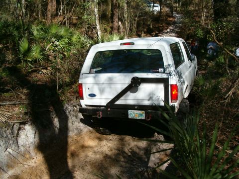 1991 Ford Bronco - A white Ford Bronco navigates a wooded trail with a spare tire mounted externally on the rear.