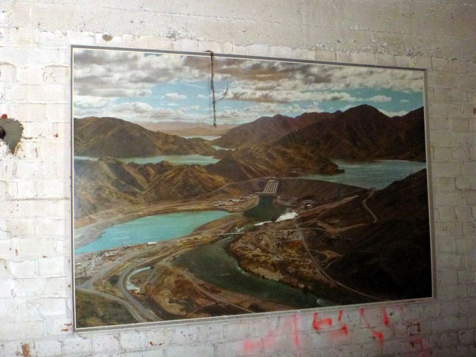Aerial landscape painting of a dam and reservoir hanging on a brick wall.