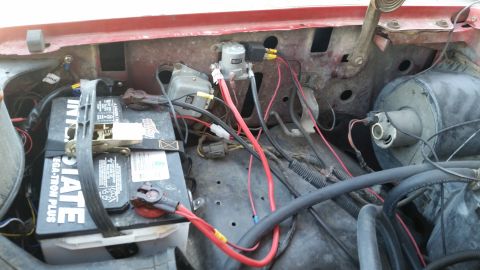 1981 Ford Bronco - The engine bay of a red vehicle shows a battery, various electrical components, and wiring. A large black battery is secured with a strap,…