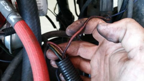 1981 Ford Bronco - A mechanic's dirty hands are holding a bundle of wires, some of which are red, yellow, green, and black, emerging from a black corrugated l…