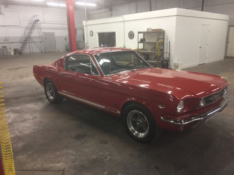 1965 Ford Mustang - A red 1965 Ford Mustang fastback is parked in a garage. The classic car is viewed from the front three-quarter angle, showcasing its chrome…