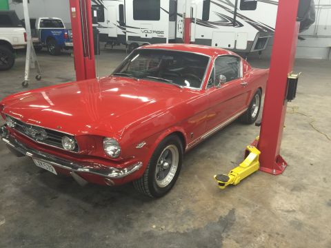 1965 Ford Mustang - A bright red classic Ford Mustang is parked on a lift in a garage. The car is a coupe with chrome accents and visible racing stripes on the…