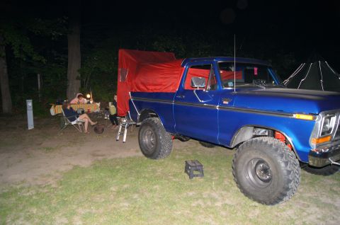 1979 Ford Bronco - A blue Ford pickup truck with a red canvas cover over the bed is parked at a campsite at night. People are sitting at a table in the backgr…