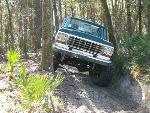 1991 Ford Bronco - A dark blue 1978 Ford Bronco navigates a challenging off-road trail through a wooded area.