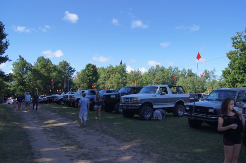 1979 Ford Bronco - A line of lifted Ford Broncos and trucks are parked on a grassy field under a blue sky with scattered clouds. Orange flags on poles are vis…