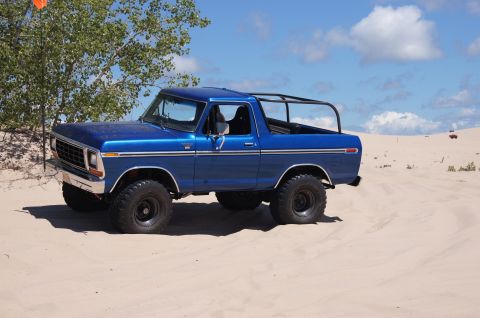 1979 Ford Bronco - A blue 1979 Ford Bronco with a roll cage sits on a sandy dune under a blue sky with scattered clouds. The vehicle is parked on a sandy surf…