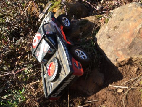 1989 Ford Bronco - An overturned remote-controlled truck with "Cummins" and "Bosch" logos is resting on its side amidst rocks and dry brush.