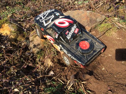 1989 Ford Bronco - A black remote-controlled monster truck with red and white decals is parked on a dirt path surrounded by dry brush and rocks. The truck has…