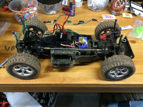 1989 Ford Bronco - A disassembled remote-controlled car chassis with its wheels, suspension, and electronic components visible on a wooden workbench.