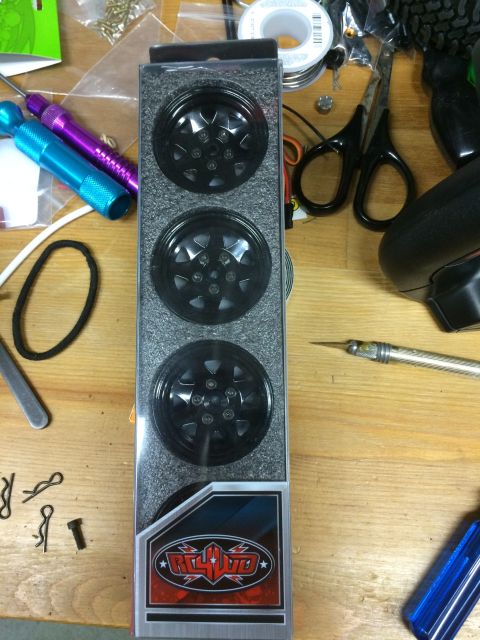1989 Ford Bronco - A package of three black RC car wheels with a "RC4WD" logo is shown on a wooden surface. Various tools and small parts are scattered around…