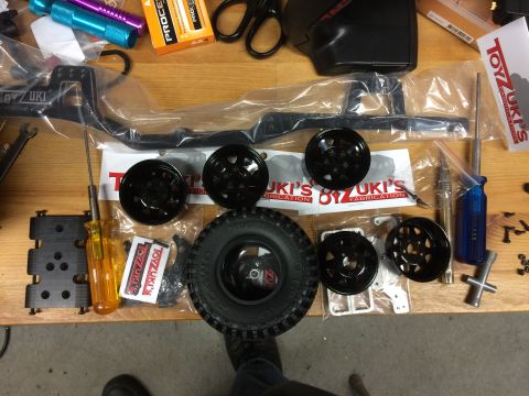 1989 Ford Bronco - A collection of RC car parts, including several black wheels, a tire, and various metal and plastic components, are laid out on a wooden su…