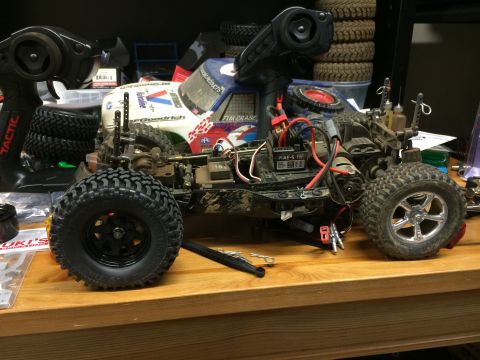 1989 Ford Bronco - A remote-controlled off-road truck with a white and blue body sits on a wooden surface, surrounded by spare tires and RC car parts. The tru…