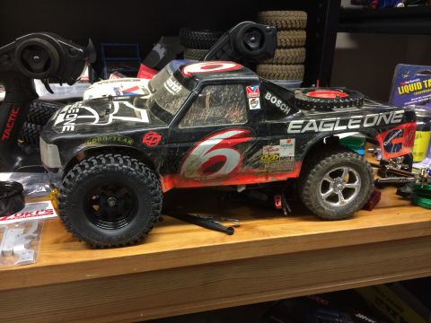 1989 Ford Bronco - A black remote-controlled monster truck with red and white racing decals sits on a wooden shelf. The truck has large knobby tires, a visibl…