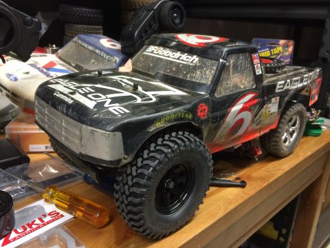 1989 Ford Bronco - A black remote-controlled pickup truck with racing decals is shown from a 3/4 front angle, resting on a wooden surface alongside other RC c…