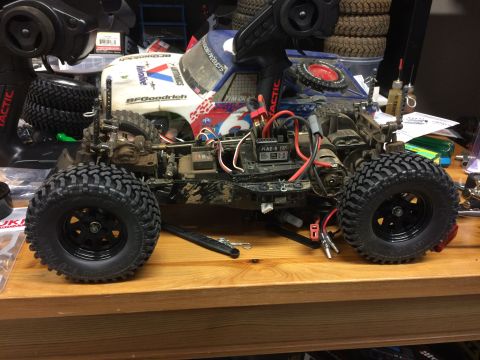 1989 Ford Bronco - A heavily modified RC truck chassis with a partially visible white and blue body sits on a wooden surface. The RC truck features large, kno…
