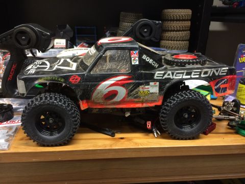 1989 Ford Bronco - A heavily modified, black remote-control monster truck with red and white racing decals sits on a wooden surface. The truck features large,…