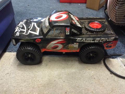 1989 Ford Bronco - A black remote-controlled monster truck with red and white racing decals sits on a carpeted floor. The truck features large, knobby tires a…