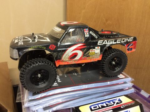 1989 Ford Bronco - A black remote-controlled monster truck with orange and white racing decals sits on top of a stack of plastic-wrapped items. The truck has…