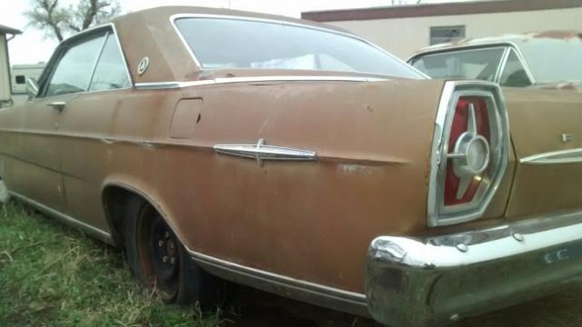 Rusty brown 1965 Ford LTD, rear passenger side view, parked in grass.