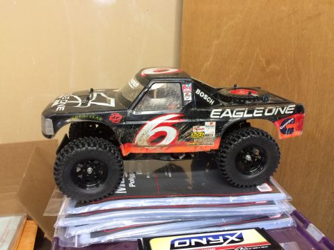 1989 Ford Bronco - A black remote-controlled monster truck with orange and white decals is shown from a side angle, resting on a stack of plastic bags. The tr…