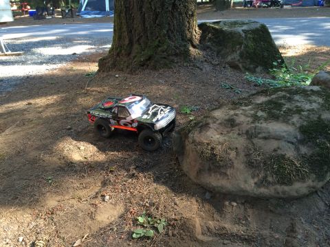 1989 Ford Bronco - A black and orange remote-controlled monster truck sits on the dirt next to a large rock and tree trunk. The truck has the text "EAGLE ONE"…