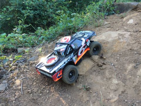 1989 Ford Bronco - A remote-controlled rock crawler truck with a black body and orange and white decals is navigating a rocky, dirt path with green foliage in…