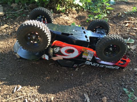 1989 Ford Bronco - An upside-down remote-controlled car with large off-road tires rests on dirt and leaves. The car has a black chassis and a red and white bo…
