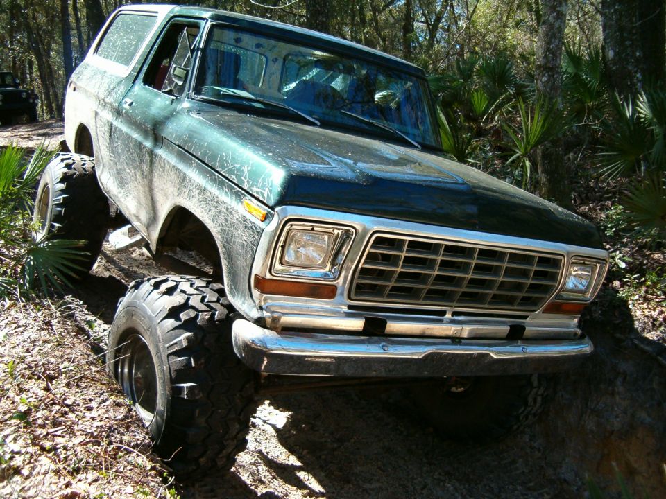 Green Ford Bronco with large tires on a dirt trail, another vehicle in background