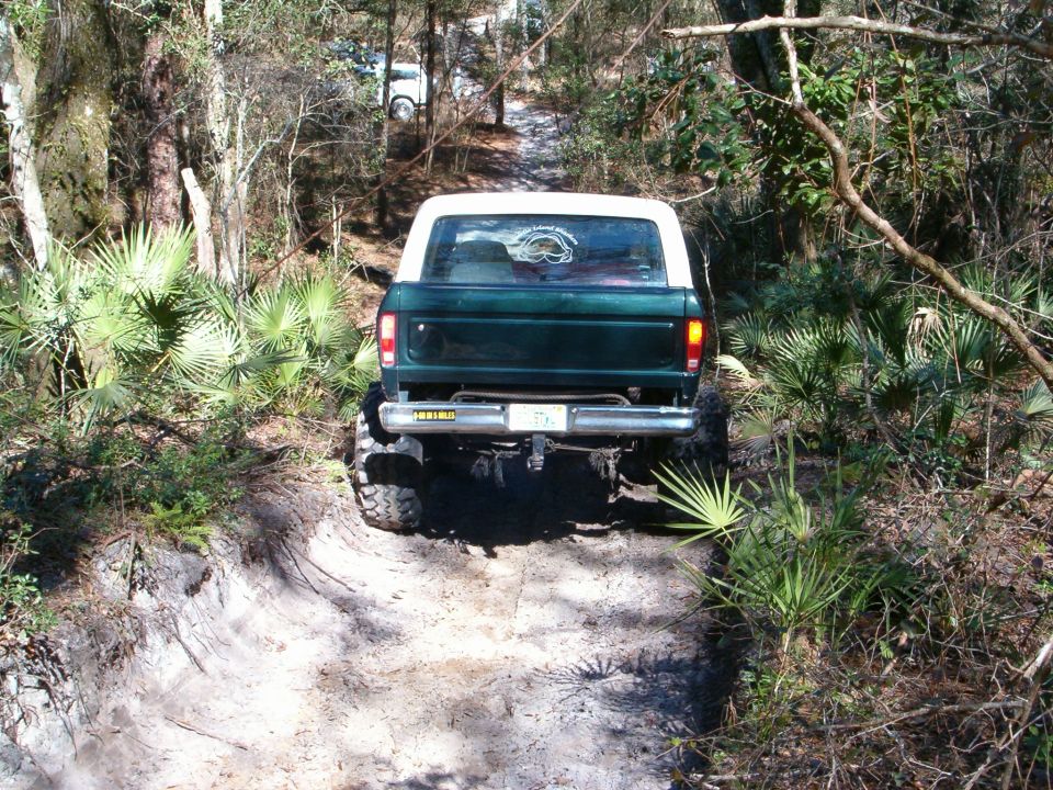 Green Ford Bronco with white top driving on a dirt path, rear view. Lifted suspension and off-road tires visible
