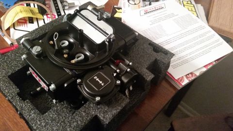 1981 Ford Bronco - A new black Quick Fuel Technology carburetor is shown packed in foam padding on a wooden table, alongside its instruction manual.