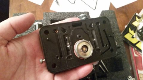 1981 Ford Bronco - A hand holds the underside of a black carburetor base plate with a brass float bowl assembly visible. The part is resting on a gray foam in…