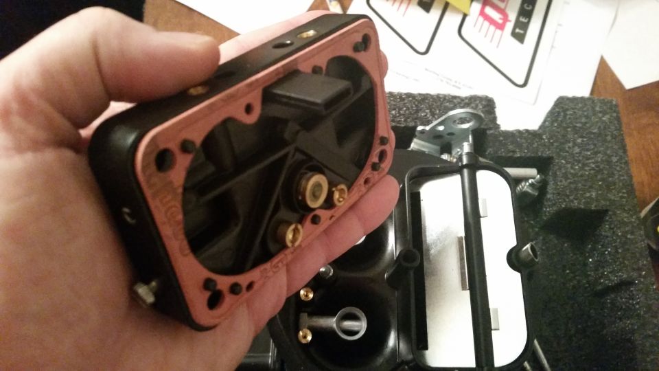 Disassembled carburetor with copper gasket held in hand, partially in a black foam box.