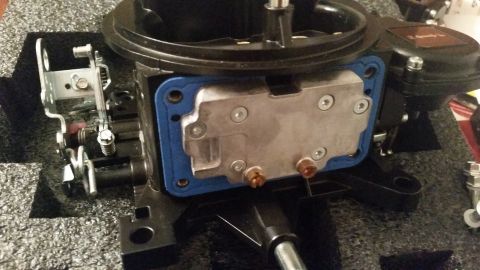 1981 Ford Bronco - A close-up view of a carburetor base plate with a blue gasket installed. Various linkage components and screws are visible on the left and…