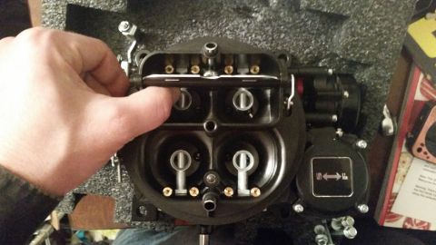 1981 Ford Bronco - A hand is shown adjusting the linkage on a black, four-barrel carburetor, which is sitting in a black foam-lined box. A red and black compo…