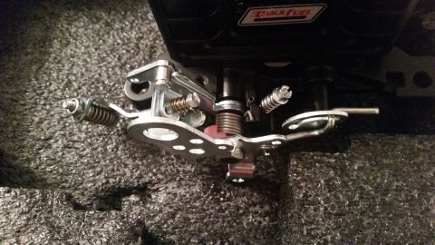 1981 Ford Bronco - A close-up view of a carburetor linkage assembly with multiple springs and metal components. A black component with a red and white "Quick…