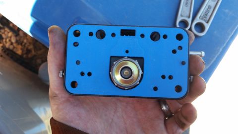 1981 Ford Bronco - A hand holds a blue gasket with a brass and silver fitting in the center. Two wrenches are visible in the background, resting on a blue sur…