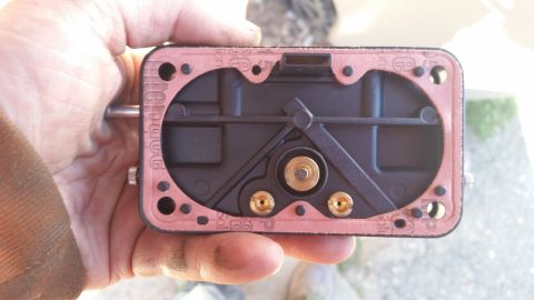 1981 Ford Bronco - A hand holds a black carburetor base with a pink gasket attached. The gasket has visible text and logos, and the carburetor base has brass…