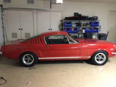 1965 Ford Mustang - A red 1965 Ford Mustang fastback is parked in a garage. The car is viewed from the side, showing its classic lines and racing stripes.