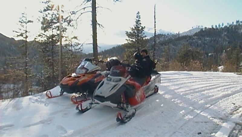 Two snowmobiles parked on a snowy hill with mountains and trees in the background.