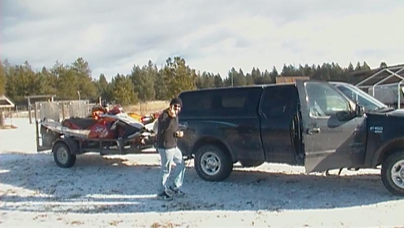 Black pickup truck with cap and trailer carrying two snowmobiles on snowy ground.