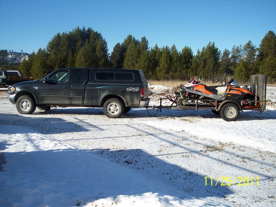 Dark pickup truck towing a trailer with two snowmobiles in a snowy landscape.