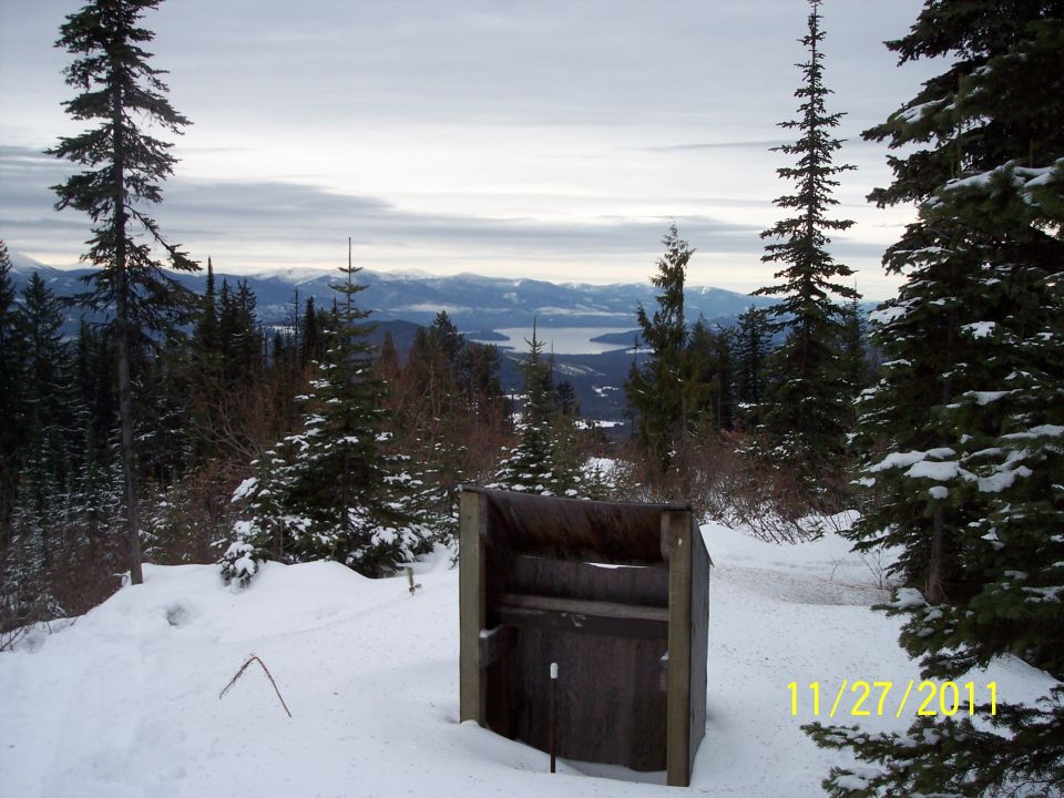 Snowy mountain landscape with a wooden outhouse structure in the foreground and a lake in the distance